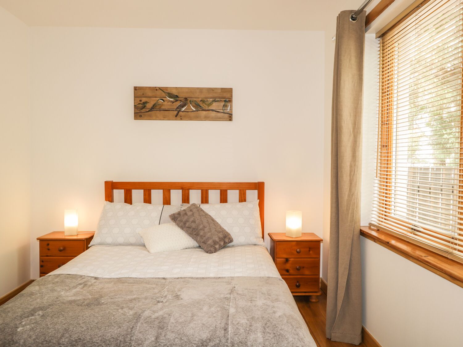 Une chambre à coucher avec un lit en bois, deux tables de chevet, une commode, une fenêtre avec des stores et un tableau accroché au mur.