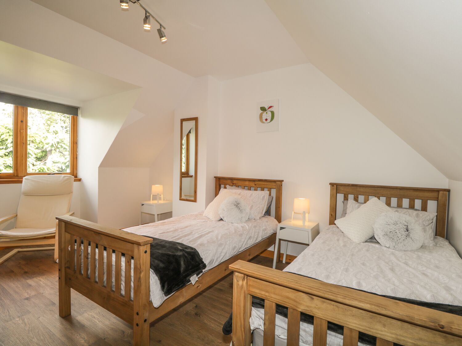 A bedroom with two wooden beds, a chair, a bedside table, a mirror, and a window with a view of greenery.
