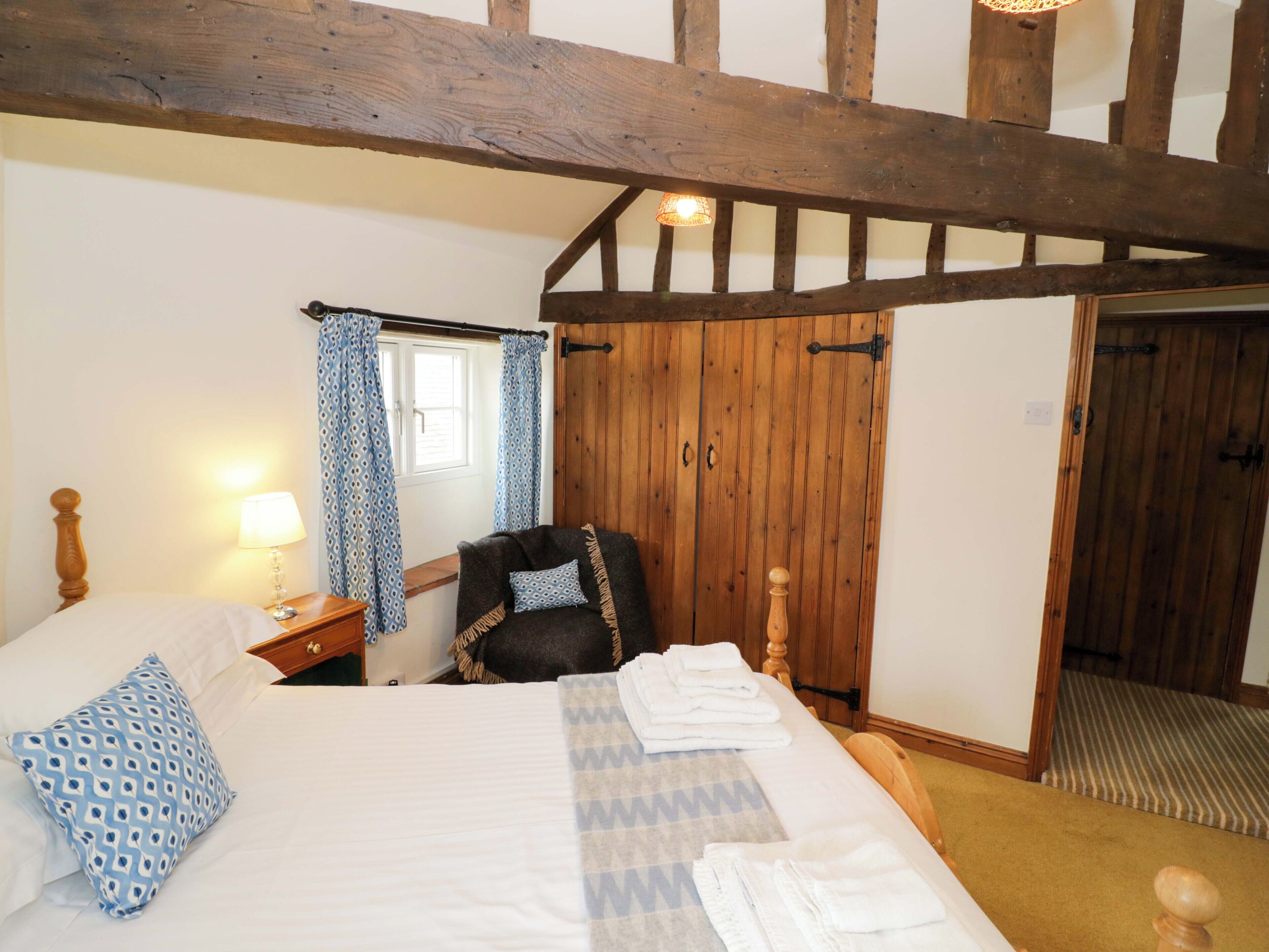 Une chambre avec un plafond en bois, un lit recouvert de draps blancs, une chaise, une table de chevet avec une lampe et une fenêtre ornée de rideaux bleus.