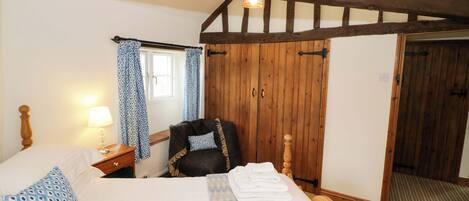 Une chambre avec un plafond en bois, un lit recouvert de draps blancs, une chaise, une table de chevet avec une lampe et une fenêtre ornée de rideaux bleus.