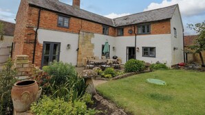 A two-story house with a brick exterior, a stone foundation, and a small garden with a lawn and potted plants. - Elmhurst Cottage (Stratford-upon-Avon)