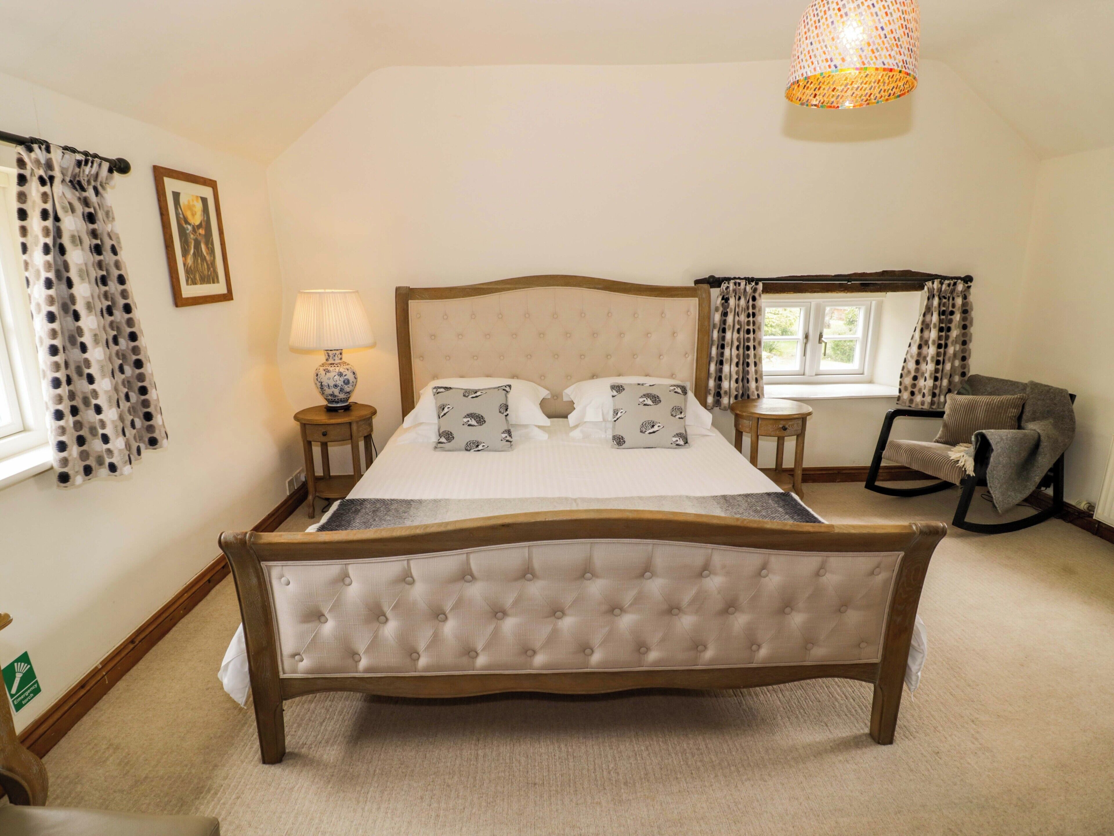 Een slaapkamer met een groot bed, een nachtkastje met lamp, een schommelstoel en een raam met gordijnen.