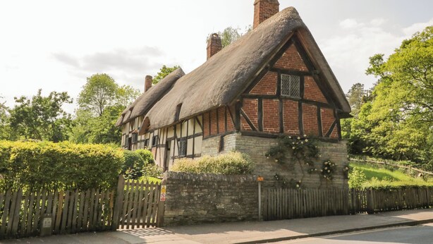A traditional half-timbered house with a thatched roof, surrounded by a wooden fence and lush greenery. - Elmhurst Cottage (Stratford-upon-Avon)