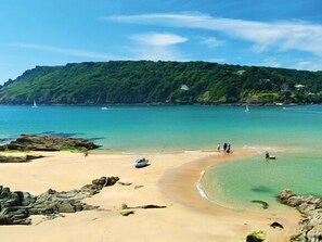 Une plage à l’eau turquoise et limpide, avec une côte rocheuse et une colline boisée en arrière-plan.
