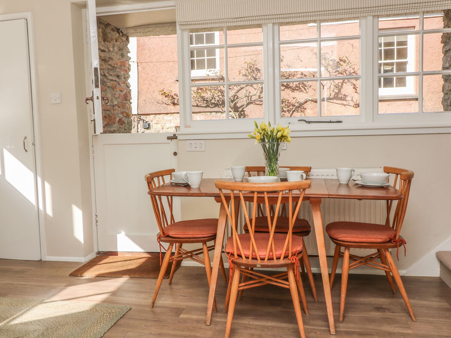 Un área de comedor con una mesa y sillas de madera, un jarrón con flores y vista a un muro de piedra y árboles por la ventana.