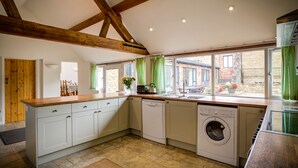 A kitchen with a wooden ceiling, white cabinets, a washing machine, and a window with green curtains. - Wytons Piece (Shipston on Stour)