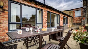 An outdoor patio with a wooden table set for dining, surrounded by brick walls and large windows. - Wytons Piece (Shipston on Stour)