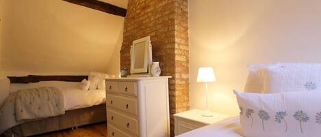 A bedroom with a brick wall, a wooden ceiling beam, a dresser, a bedside table with a lamp, and two beds.