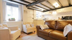 A cozy living room with a brown leather sofa and two white armchairs, a wooden floor, and a kitchen with white cabinets and a brick backsplash. - One Masons Court (Stratford-upon-Avon)