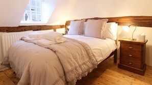 A bedroom with a sloped ceiling, a wooden headboard, a bed with white linens, a bedside lamp, and a wooden nightstand. - One Masons Court (Stratford-upon-Avon)