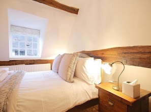 A bedroom with a wooden headboard, a bedside table with a lamp and tissues, and a window with blinds. - One Masons Court (Stratford-upon-Avon)