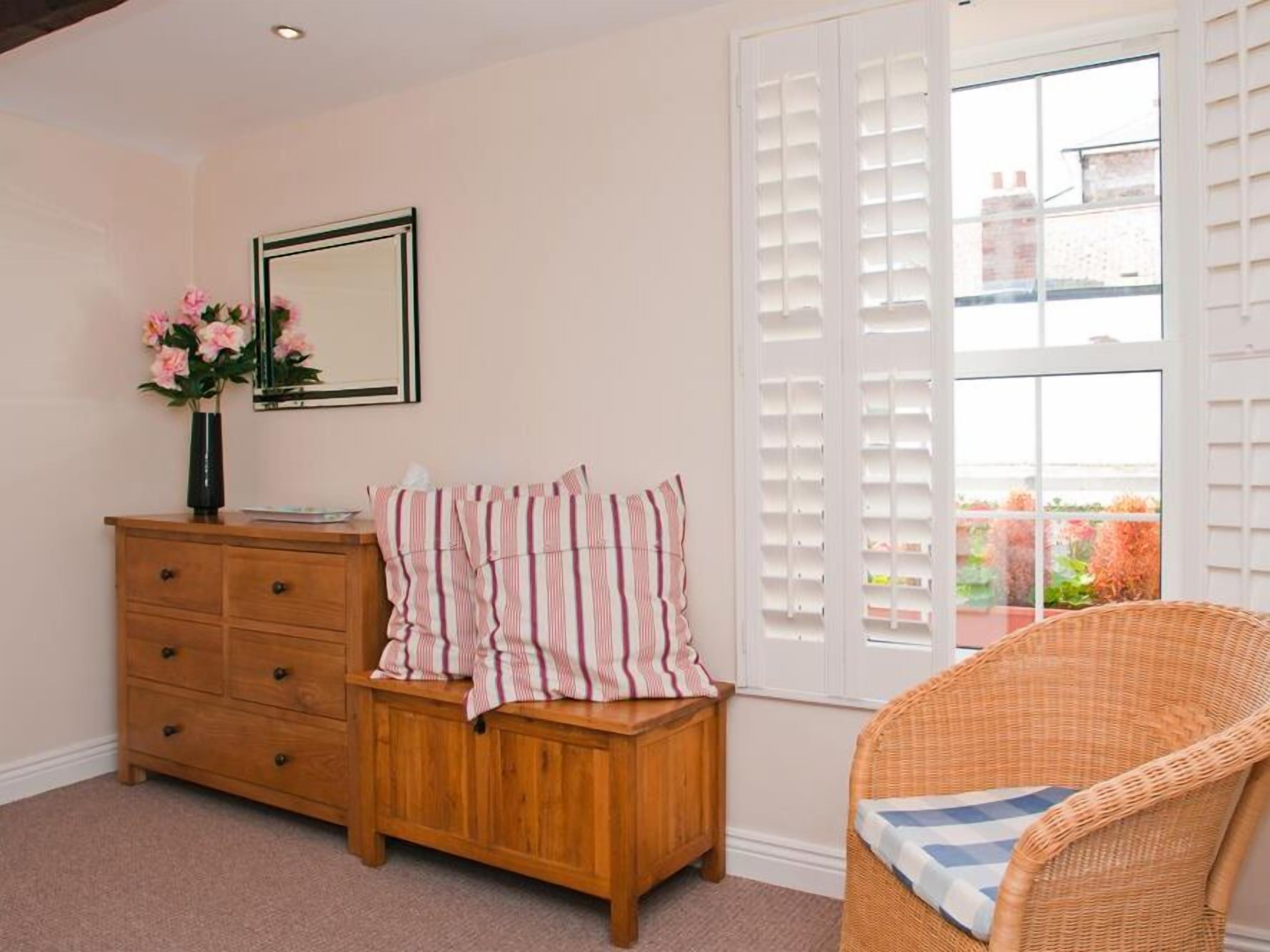 A room with a wooden dresser, a wicker chair, a window with white shutters, and a framed mirror on the wall.