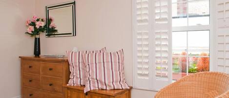 A room with a wooden dresser, a wicker chair, a window with white shutters, and a framed mirror on the wall.