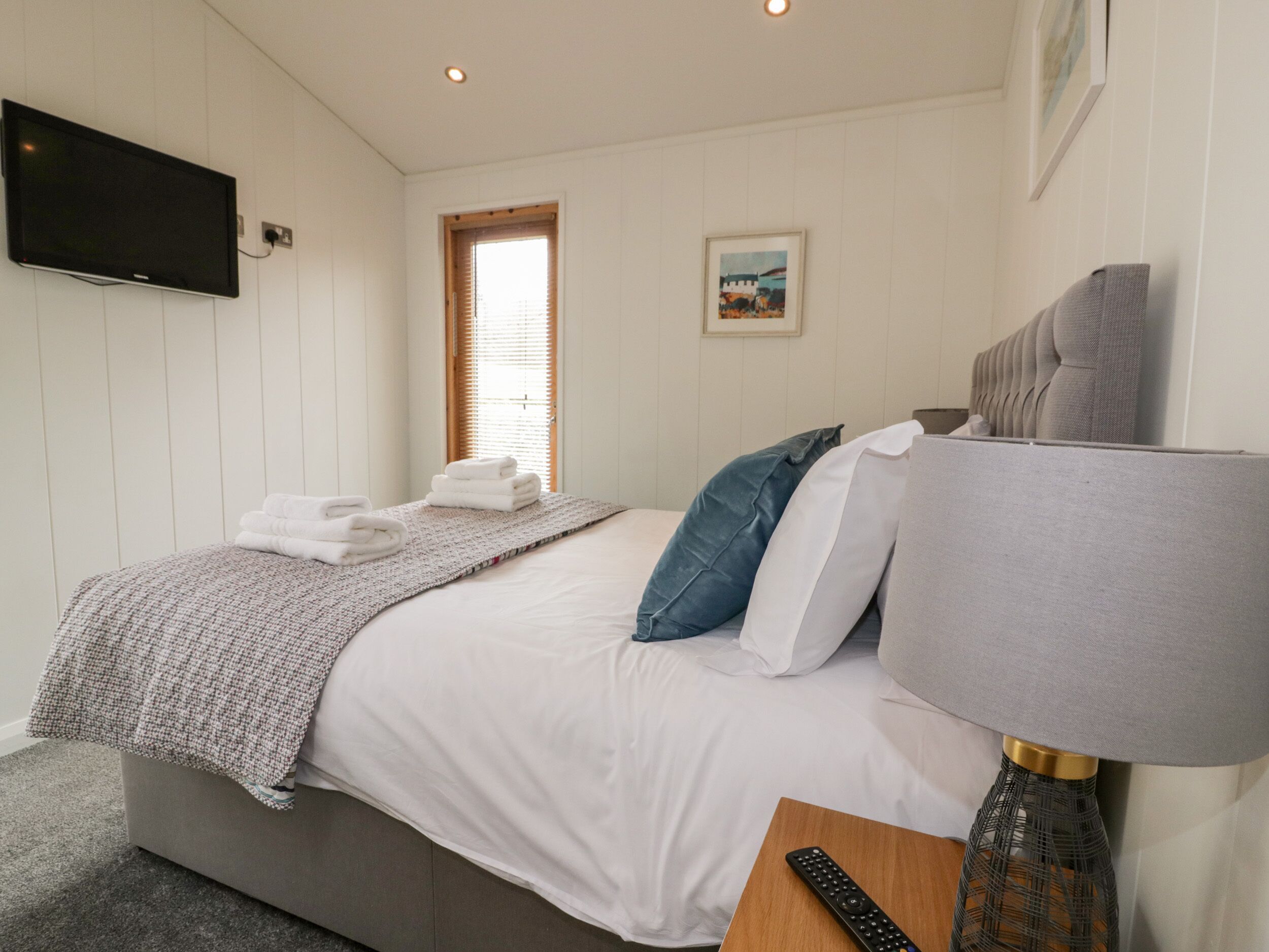 A neatly made bed with a grey headboard, a bedside table with a remote control, and a wall-mounted TV.