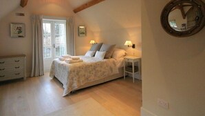 A bedroom with a bed, bedside lamps, a mirror, a dresser, and a window with curtains. - Hillside Cottage (Burford)