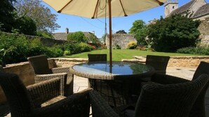 A round outdoor table with wicker chairs under a large umbrella, set against a backdrop of stone walls and greenery. - Hillside Cottage (Burford)