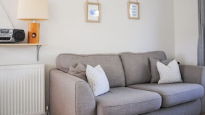 A neutral-toned living room with a grey sofa, two framed pictures on the wall, a wooden side table with a lamp, and a radiator. - Bankside (Kingsbridge)