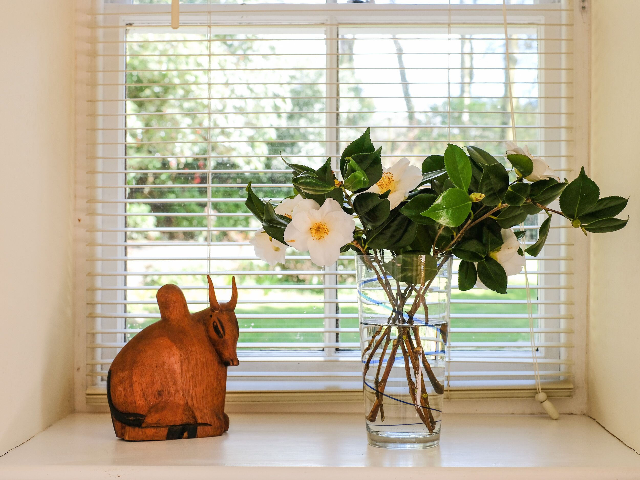 Una ventana con persianas blancas, vistas despejadas a follaje y un florero de vidrio con ramas y flores blancas en el alféizar.