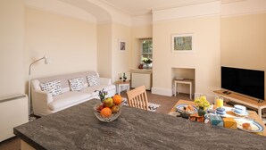 A modern living room with a white sofa, a dining table with breakfast items, and a TV on a stand. - The Patio Flat (Bodmin)