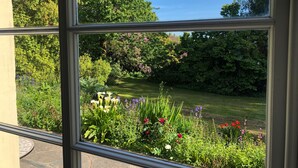 A view through a window of a garden with various plants and flowers, a lawn, and trees. - The Patio Flat (Bodmin)