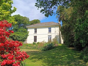 Un bâtiment à deux étages, doté d’une façade symétrique, entouré d’une végétation luxuriante et de plantes en fleurs.
