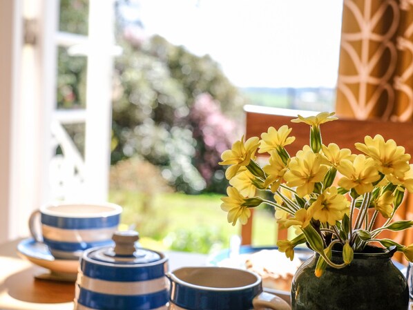 A table with a vase of yellow flowers, blue and white ceramic containers, and a window with a view of greenery. - The Patio Flat (Bodmin)