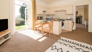 A spacious living room with a television, a dining area with a table and chairs, a kitchen with modern appliances, and a view of the garden through the open door. - The Patio Flat (Bodmin)