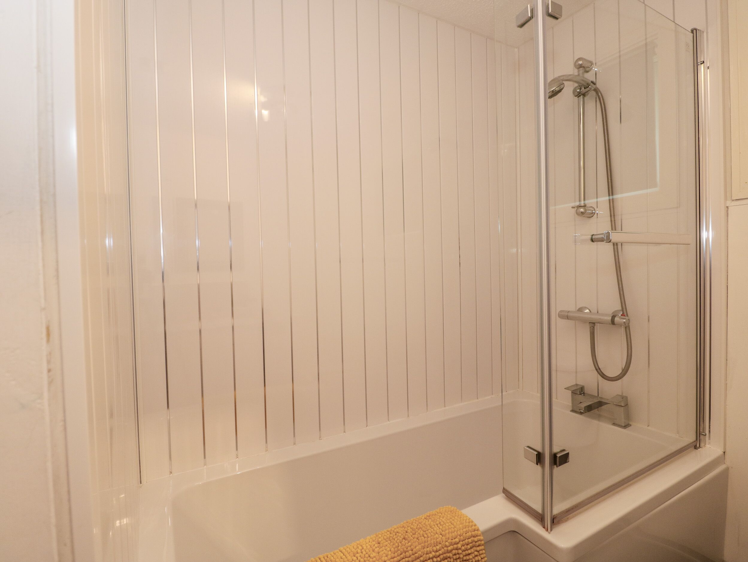 A modern bathroom with a glass-enclosed shower, white tiled walls, and a yellow bath mat.