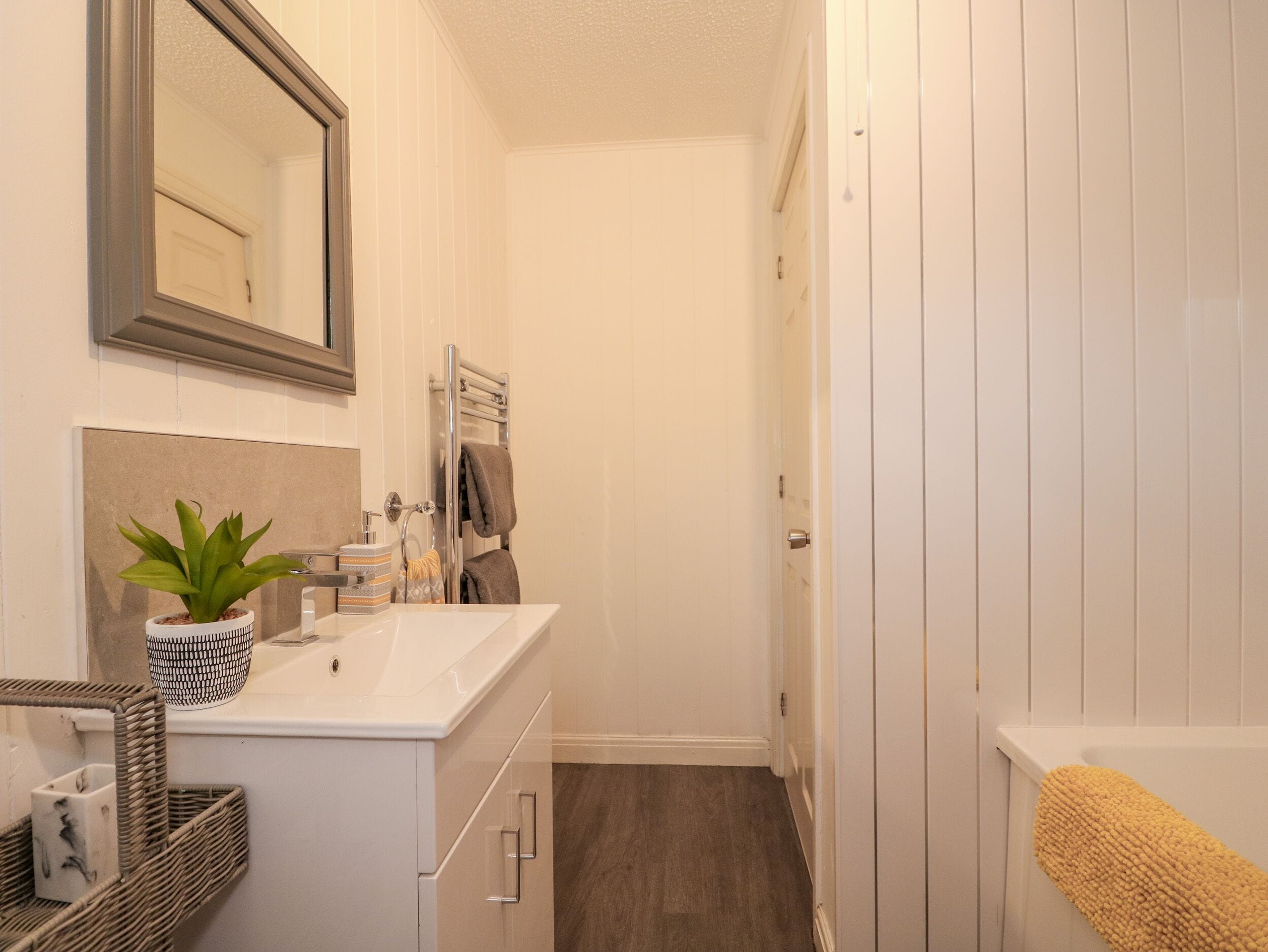 A narrow bathroom with a white sink, a mirror, a towel rack, a potted plant, and a yellow bath mat.