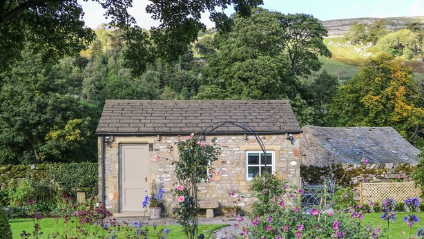 A stone cottage with a tiled roof, surrounded by a well-maintained garden with various flowers and a stone wall. - The Bothy (Skipton)