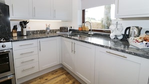 A modern kitchen with white cabinets, a marble countertop, a built-in oven, and a window with a view of the outdoors. - Garth Cottage (Denbigh)