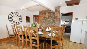 A dining area with a round table set for six, wooden chairs, a large wall clock, and a brick wall. - Garth Cottage (Denbigh)