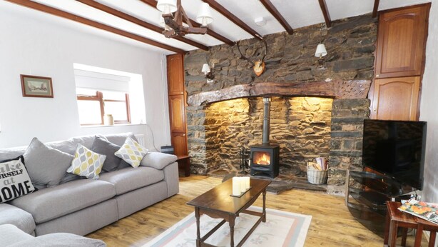 A cozy living room with a stone fireplace, a grey sofa, a wooden coffee table, and a television. - Garth Cottage (Denbigh)