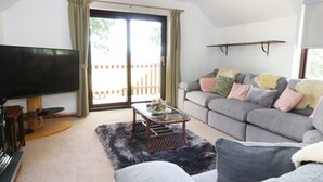 A living room with a flat-screen TV, a grey sofa, a coffee table, and a large sliding glass door leading to an outdoor area. - Garth Cottage (Denbigh)