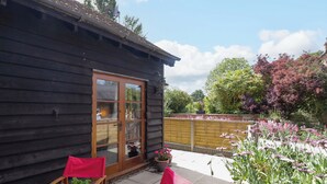 Eine Holzhütte mit einer Schiebetür, einer Terrasse mit zwei Holzstühlen und einem Tisch sowie ein Garten mit verschiedenen Pflanzen und Blumen.