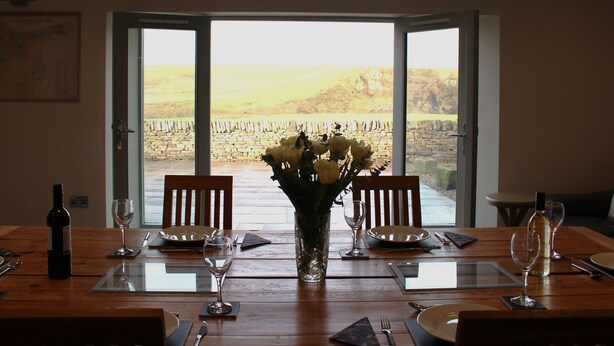 A dining table set for four with a vase of flowers, two chairs, and a view of a stone wall and fields through a glass door.