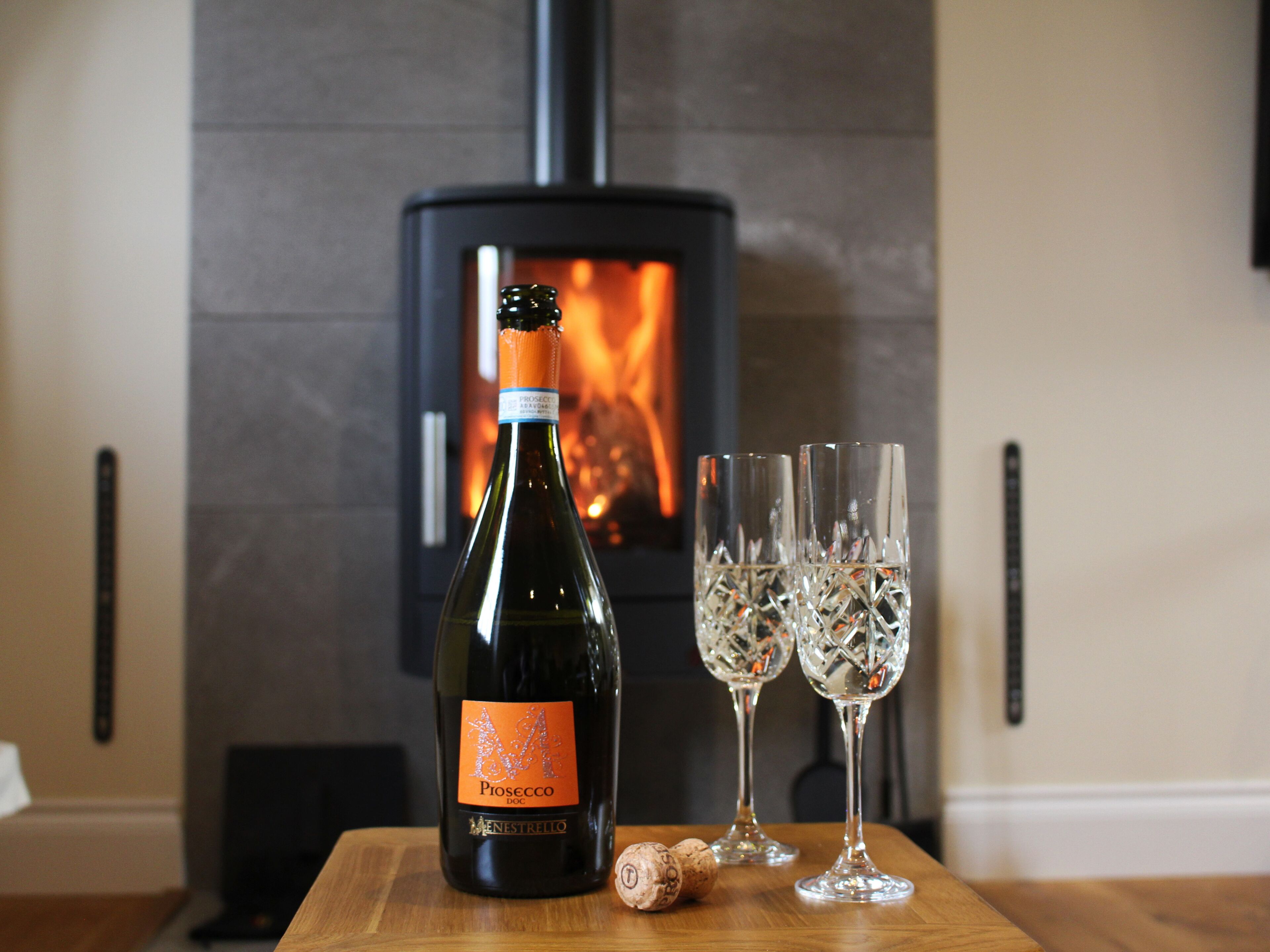 A bottle of wine, two glasses, and a cork on a wooden table with a fireplace in the background.