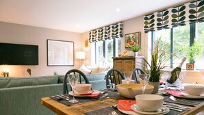 A dining area with a wooden table set for two, a green sofa in the background, and a window with patterned curtains. - Sidings at Brunel Two (Lostwithiel)
