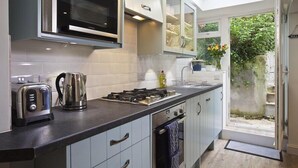 A modern kitchen with a built-in oven, microwave, and gas stove. There is a window providing natural light and a view of the garden outside. - Crab Cottage (Salcombe)