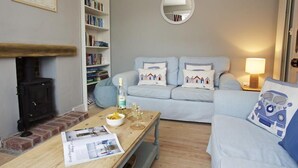 A cozy living room with a fireplace, a sofa set, a coffee table with a newspaper, and a bookshelf. - Crab Cottage (Salcombe)