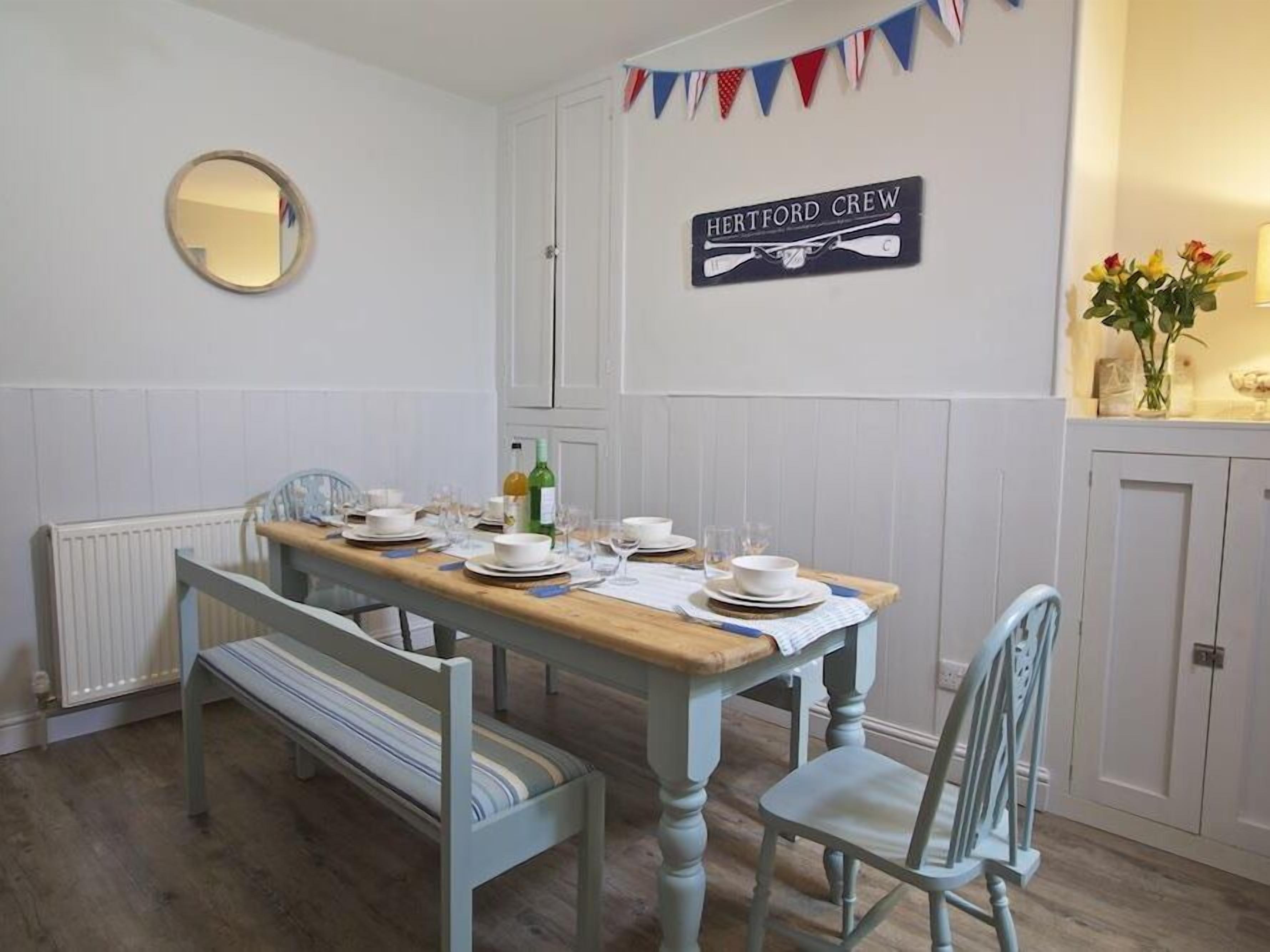 A dining room with a wooden table set for four, a mirror on the wall, and a sign reading "HERTFORD CREW".