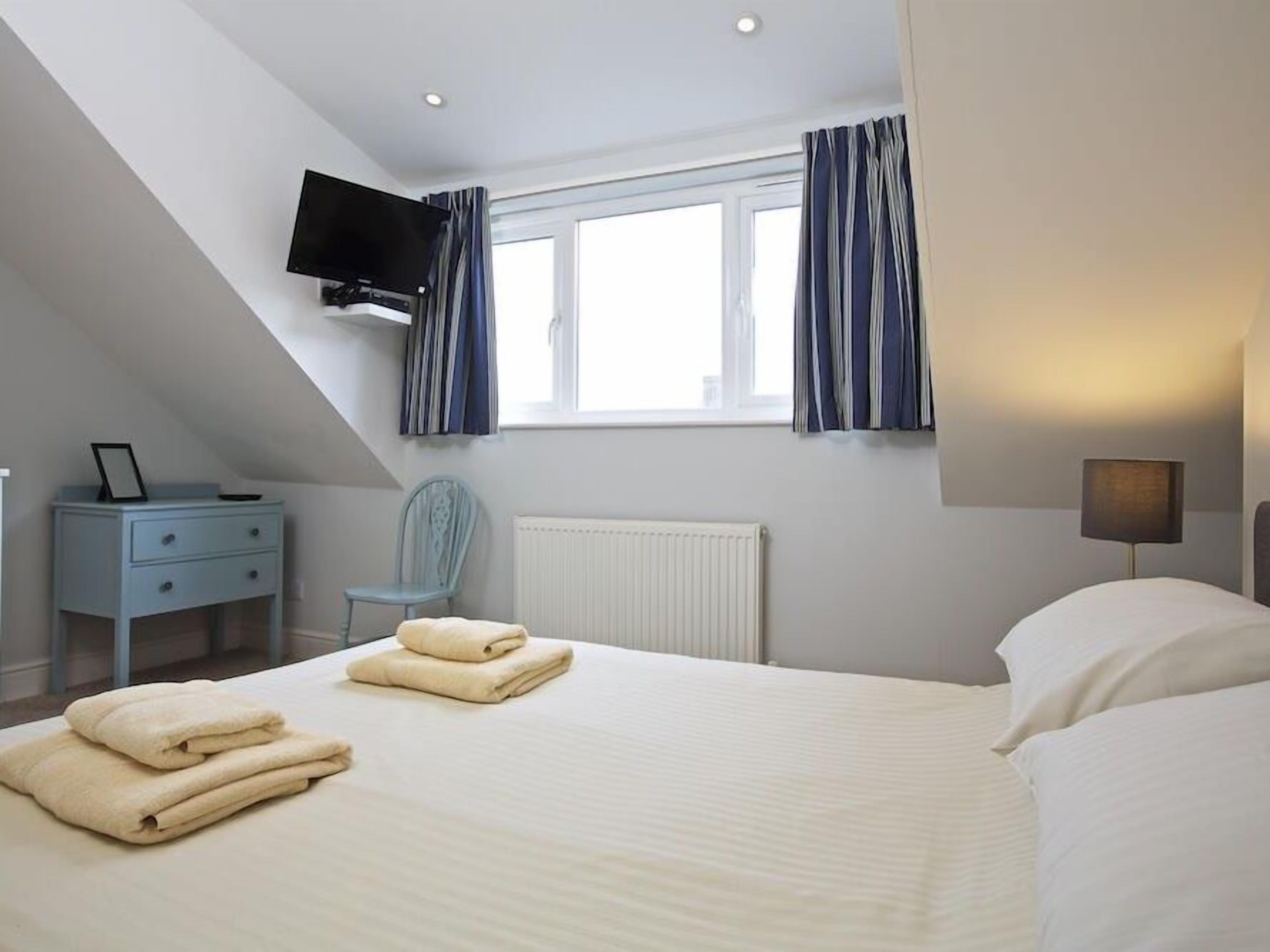 Une chambre à coucher avec un lit, un téléviseur fixé au mur, une chaise, une table de chevet et une fenêtre avec des rideaux.