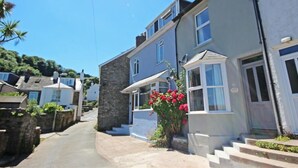 A narrow street lined with two-story houses with stone and plaster exteriors, a clear blue sky, and a small garden with red flowers in front of one house. - Crab Cottage (Salcombe)