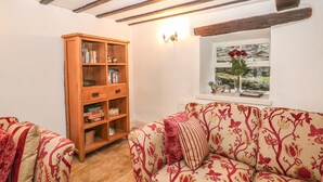 A cozy living room with a wooden bookshelf, a patterned sofa, and a window with a view of a rocky landscape. - Laburnum Cottage (Hope Valley)