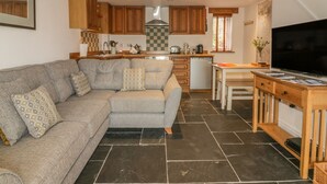 A modern living room with a grey sofa, wooden cabinets, a kitchen area with a stove, and a TV on a wooden stand. - Crift Farm Cottage (Bodmin)