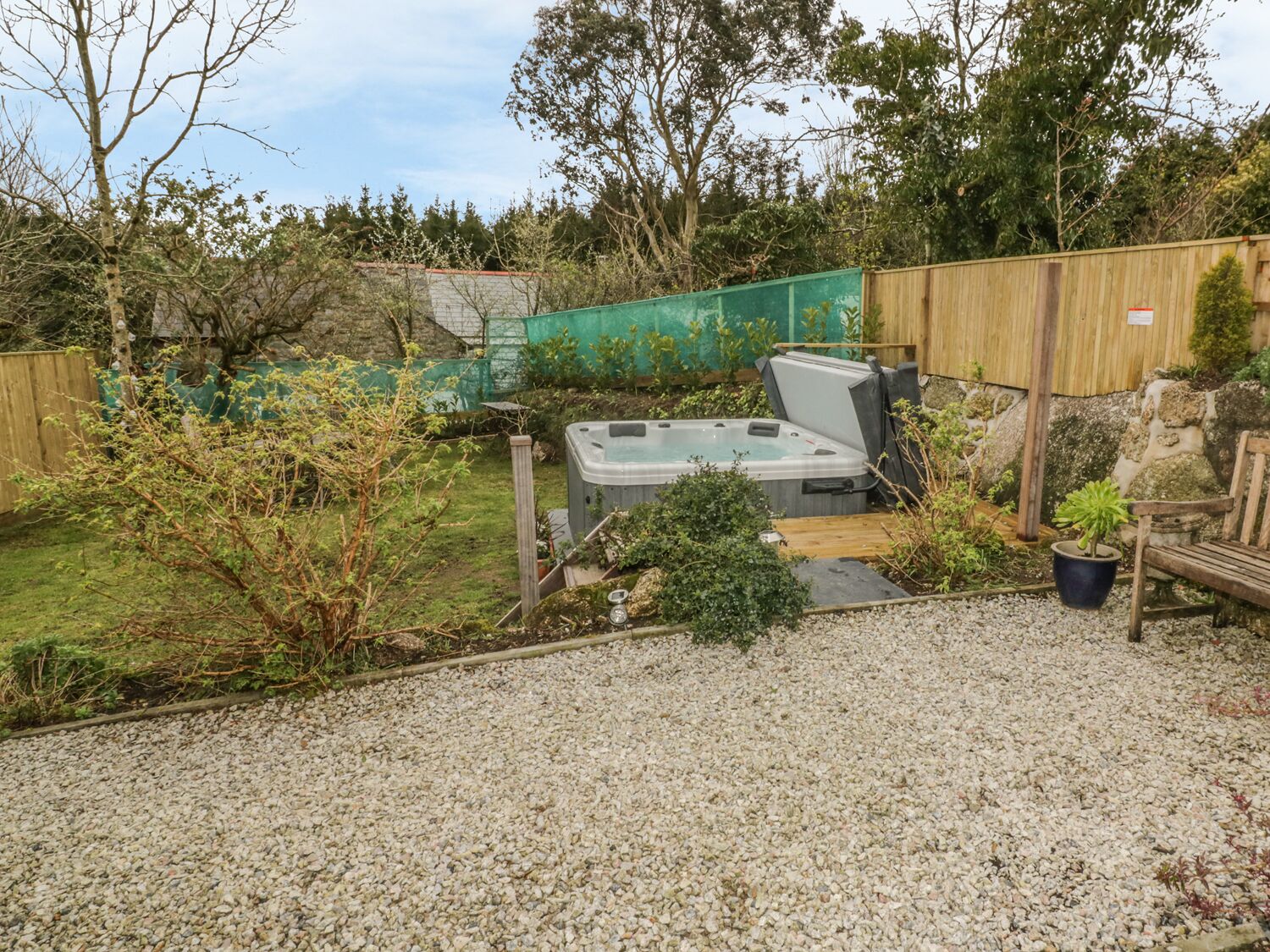 A hot tub in a garden with a wooden fence, gravel ground, and various plants.