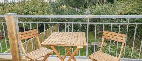 A wooden deck with two wooden chairs and a table, surrounded by a metal railing, overlooking a garden with various plants and trees.