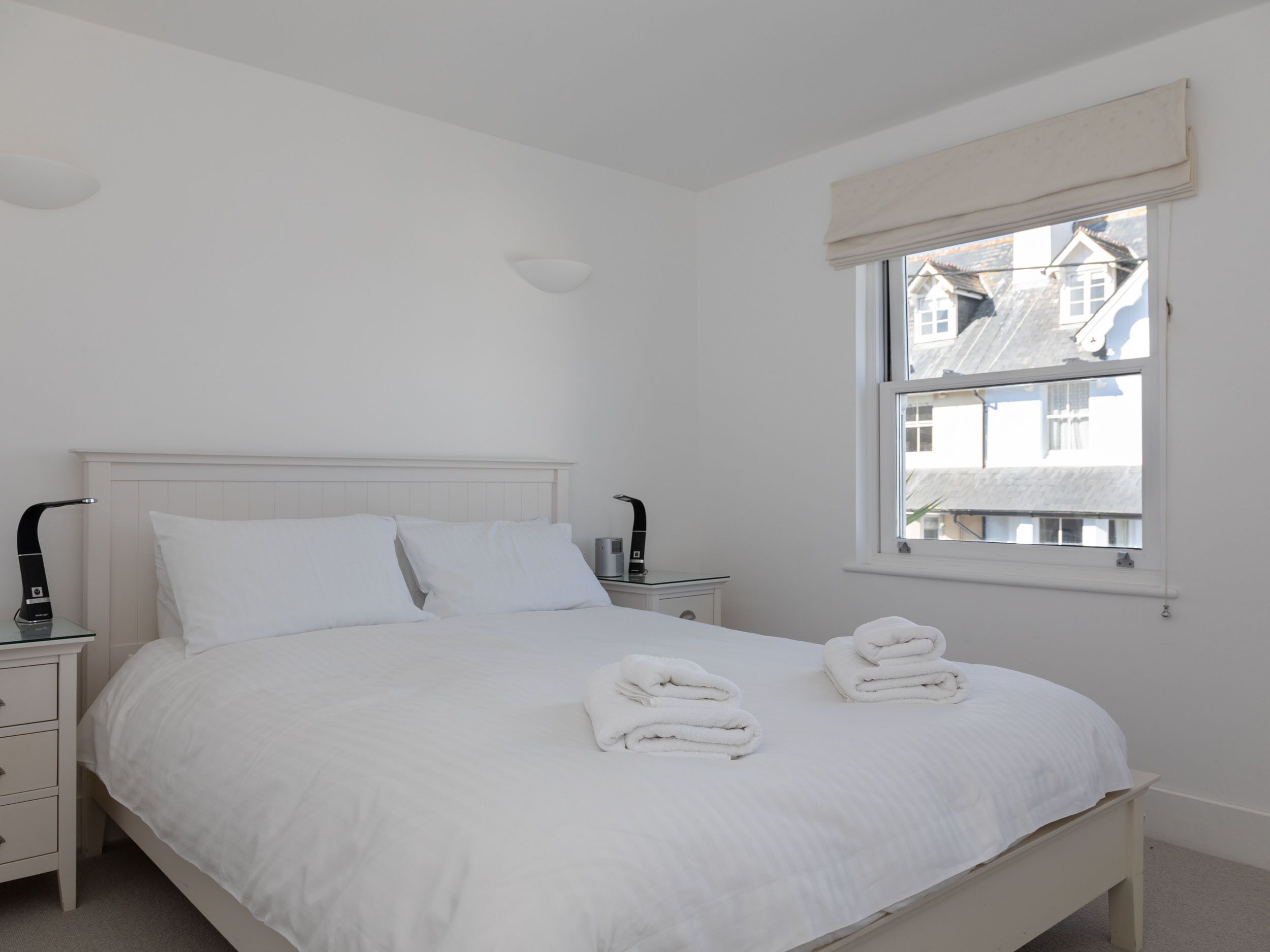A white bedroom with a bed, two bedside tables, and a window with a view of buildings.