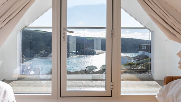 A room with a large glass door leading to a balcony with a view of a lake and hills. - 4 Blue View (Salcombe)