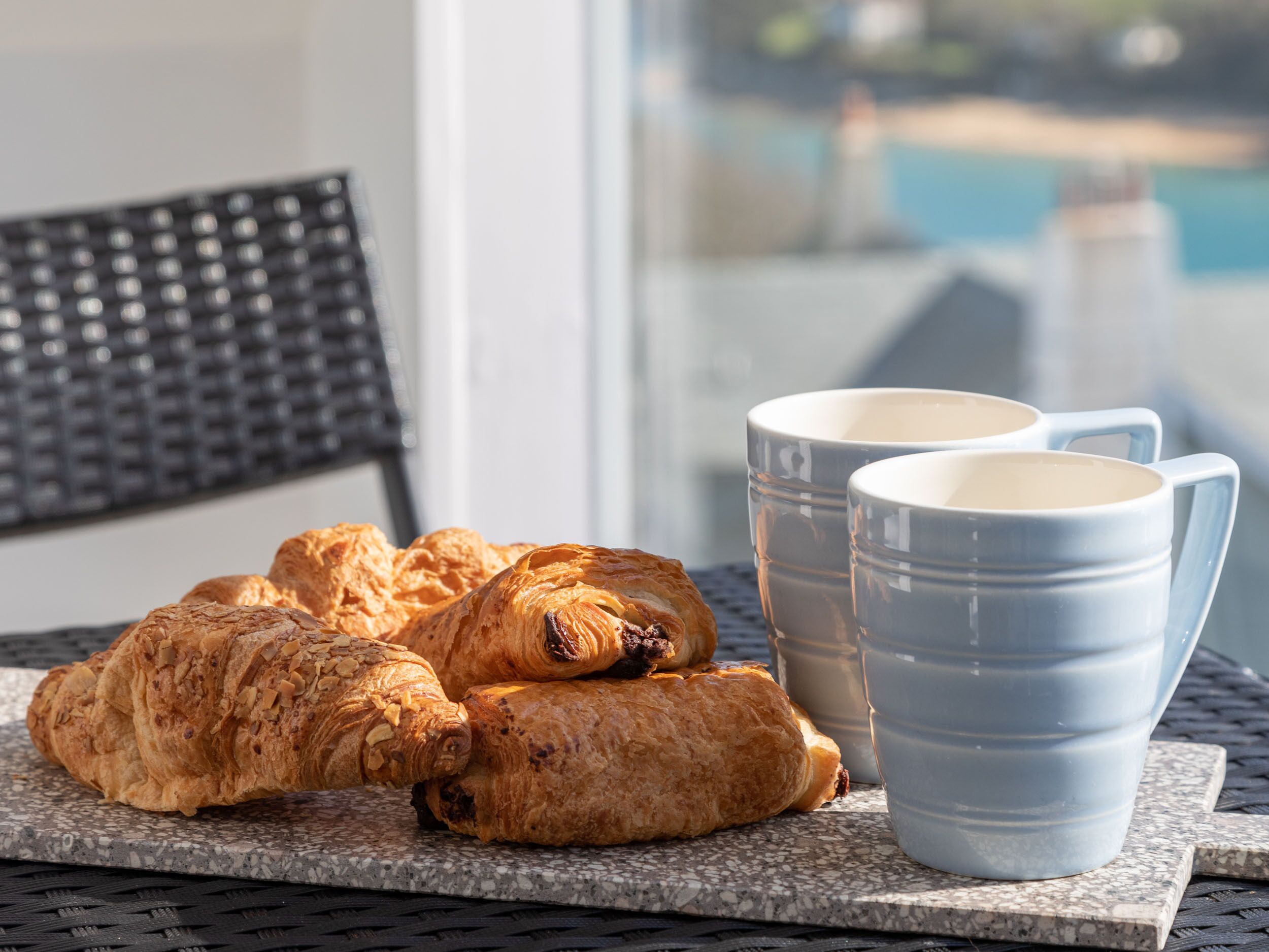 Ein Tisch mit Croissants und zwei blauen Keramiktassen, auf einem Balkon mit einem Stuhl und Blick auf einen Pool.